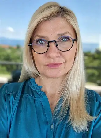 Arin portret sa naočalama u plavoj bluzi ispred prirodne pozadine.
